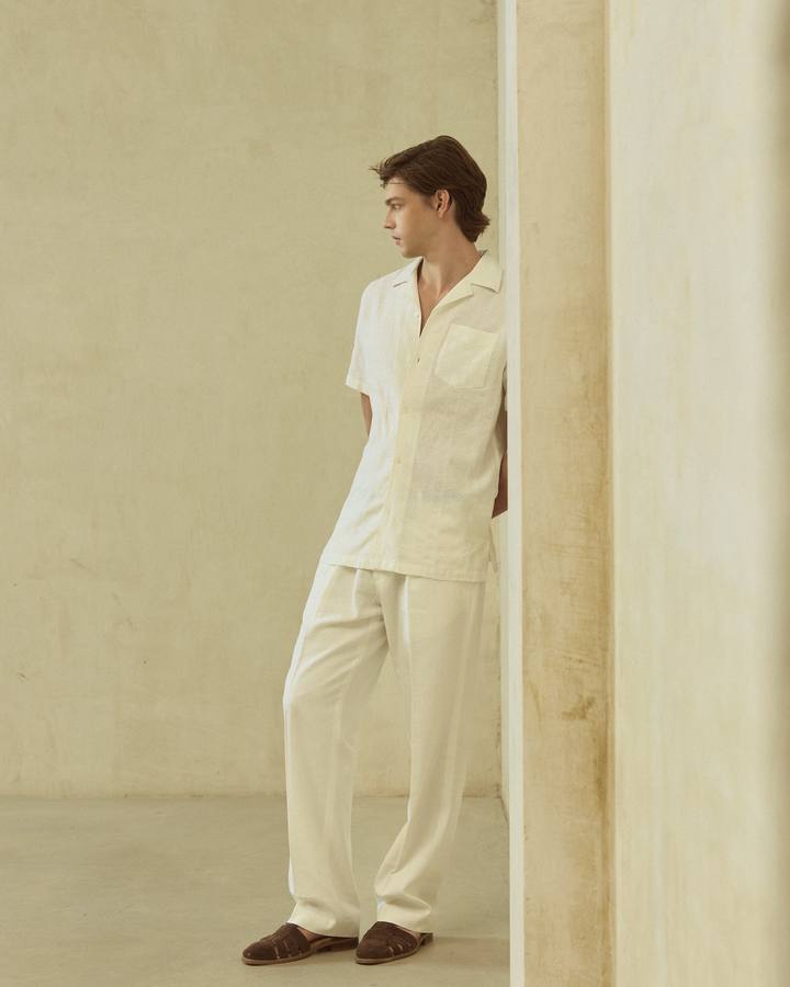 Modelo con camisa de lino manga corta blanco roto de PuroEgo y pantalón claro, de cuerpo entero, apoyado en un muro claro.