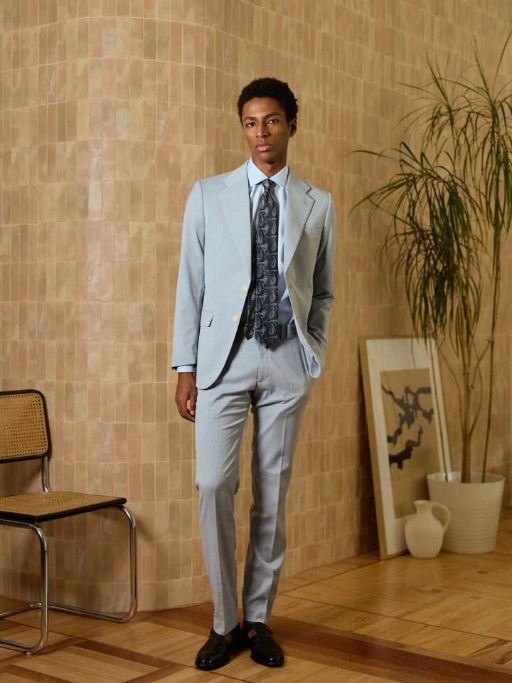 Traje estructura celeste de PuroEgo de cuerpo entero, con camisa celeste, corbata y zapatos negros, junto a un muro de azulejos.