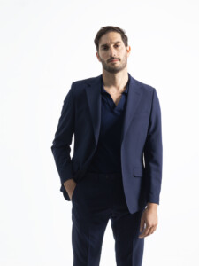 Traje 2 piezas azul marino en modelo de frente, chaqueta abierta de dos botones sobre polo azul y una mano en el bolsillo del pantalón.