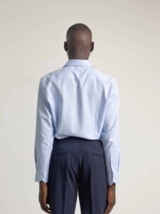 Camisa celeste PuroEgo vista de espalda sobre modelo, se aprecia la caída de la espalda y los puños abotonados junto a un pantalón azul marino.