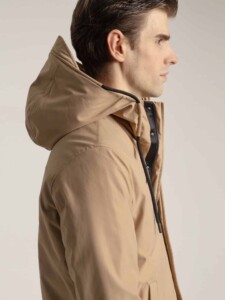Detalle en tres cuartos del abrigo beige PuroEgo: capucha armada, cierre negro con broches y cordón de ajuste sobre el hombro.