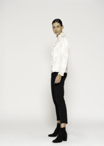 Vista de perfil lateral de blusa beige satinada, destacando cuello alto con volado y botones frontales, combinada con pantalón negro