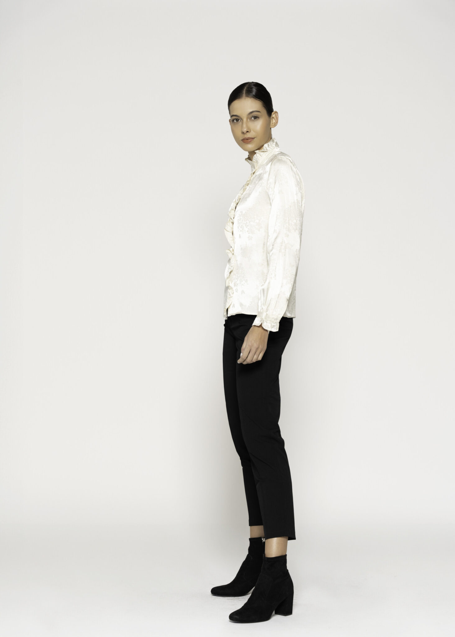 Vista de perfil lateral de blusa beige satinada, destacando cuello alto con volado y botones frontales, combinada con pantalón negro