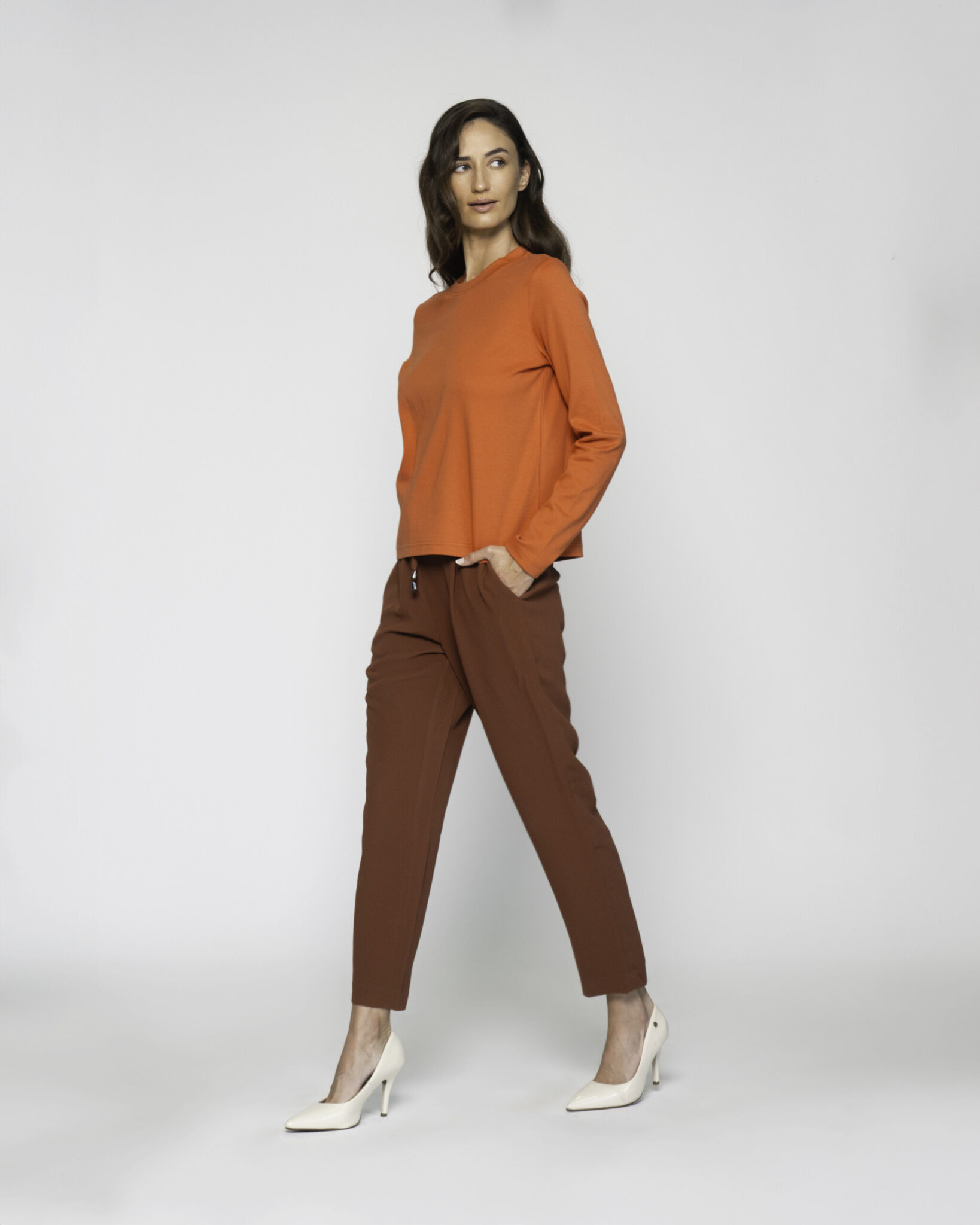 Vista lateral de camiseta naranja manga larga con cuello redondo, combinada con pantalón marrón y stilettos blancos