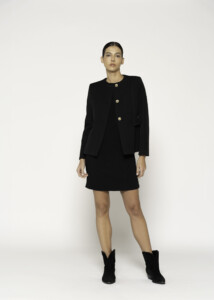 Vista completa frontal de chaqueta negra con tres botones dorados al centro, cuello redondo y corte recto, combinada con falda corta negra y botines, modelo Formentera