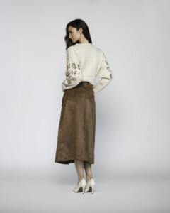 Vista de espalda de falda midi camel efecto ante con abertura trasera, combinada con suéter beige con letras modelo Formentera