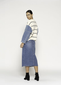 Vista lateral de tres cuartos desde atrás del sweater a rayas con mangas de mezclilla azul, mostrando la falda midi denim y botines negros