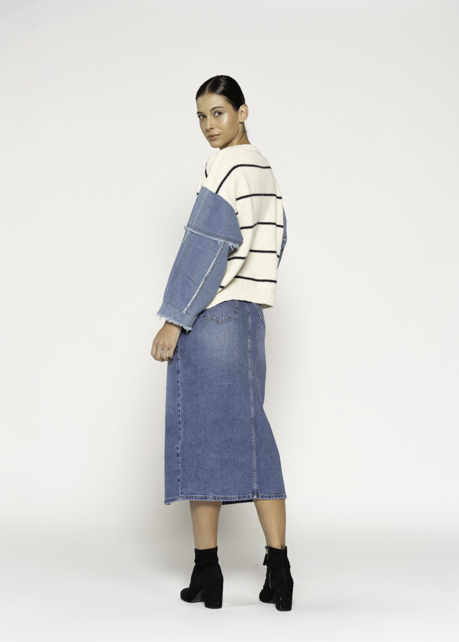 Vista lateral de tres cuartos desde atrás del sweater a rayas con mangas de mezclilla azul, mostrando la falda midi denim y botines negros
