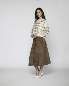 Vista lateral completa de sweater beige con letras doradas repetidas, combinado con falda midi café abotonada y stilettos blancos