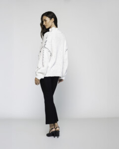 Vista lateral de sweater blanco con cuello alto, tejido grueso y líneas negras diagonales, combinado con pantalón negro
