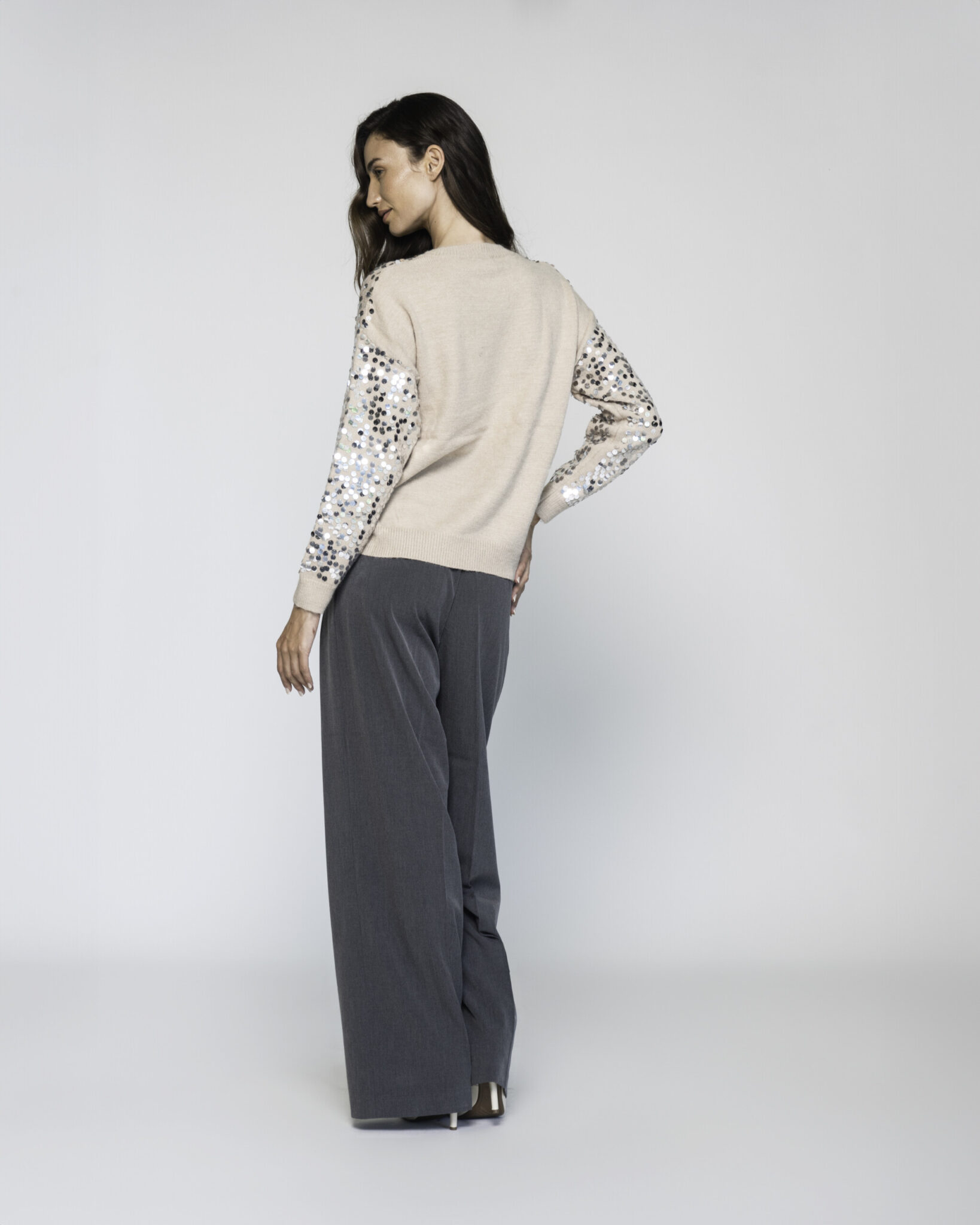 Vista de espalda del sweater beige claro, mangas con lentejuelas plateadas y negras, puños de tejido canalé, pantalón gris ancho