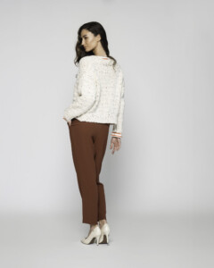 Vista lateral de sweater tejido crudo con textura moteada y detalle de puños con franjas naranjas, combinado con pantalón marrón