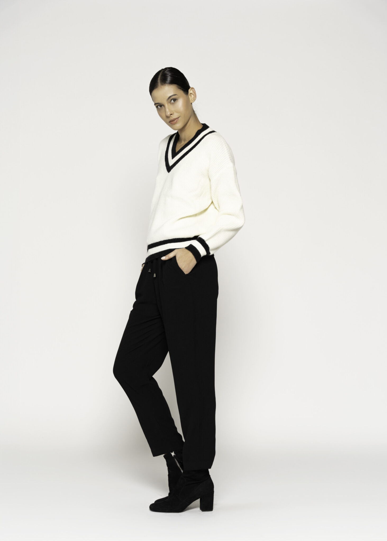 Vista lateral de tres cuartos mostrando sweater tejido crudo con ribetes negros en cuello V y puños, combinado con pantalón negro