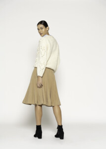 Vista de tres cuartos trasera del sweater tejido crudo con trenzas en relieve y falda beige plisada, modelo Formentera