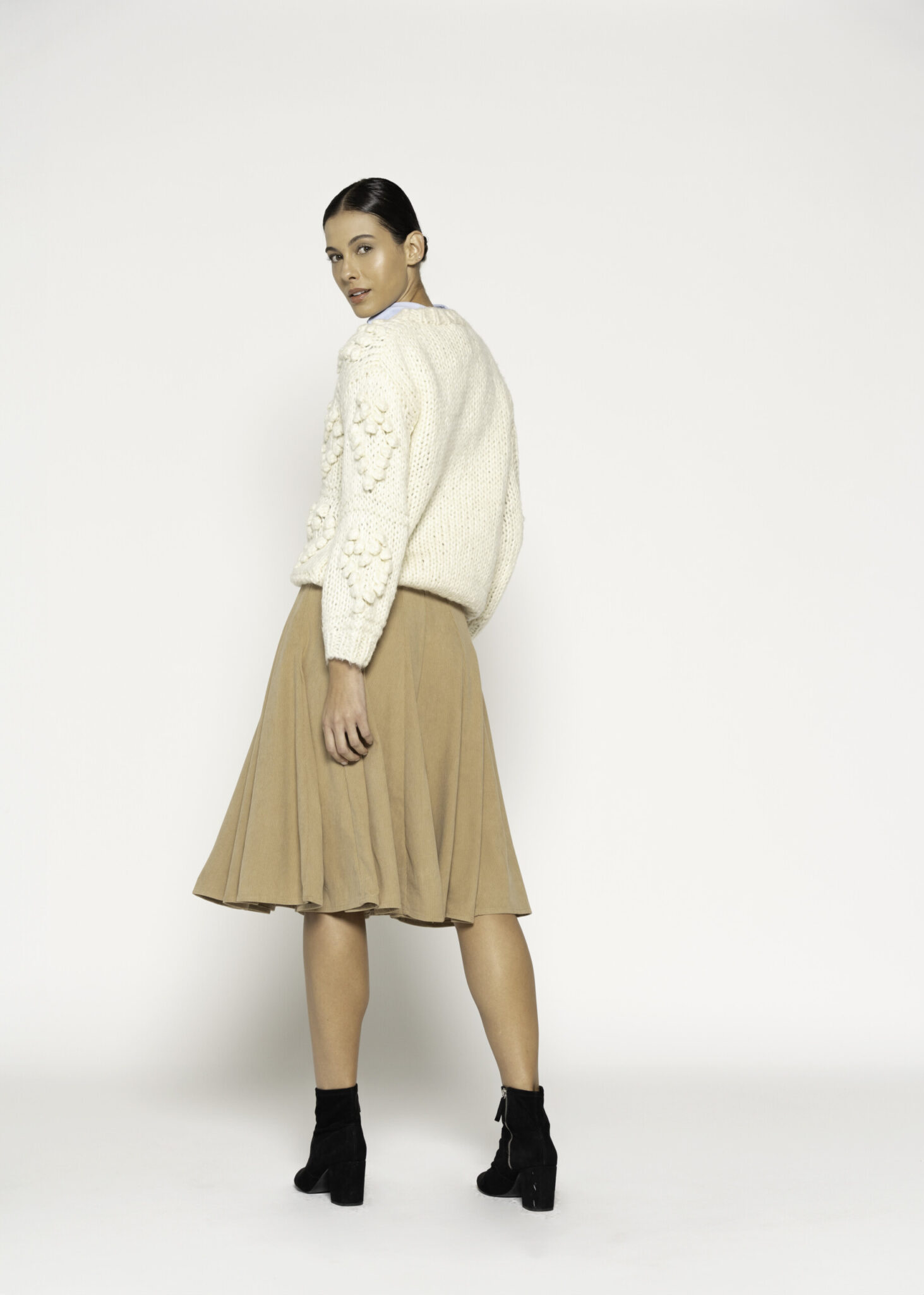 Vista de tres cuartos trasera del sweater tejido crudo con trenzas en relieve y falda beige plisada, modelo Formentera