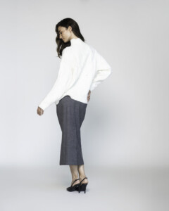 Vista lateral de sweater blanco de cuello alto y mangas largas, se aprecia el corte relajado y la caída de la prenda sobre pantalón gris