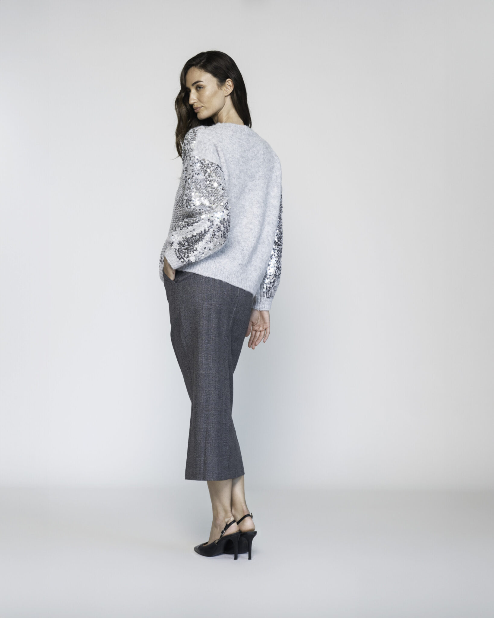 Vista lateral de sweater tejido gris claro con mangas cubiertas de lentejuelas plateadas, combinado con pantalón oscuro