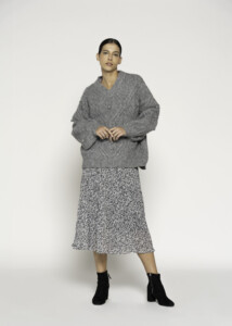 Vista completa de cuerpo entero, sweater gris oversized con cuello en V y textura trenzada, combinado con falda plisada estampada y botines negros, modelo Formentera