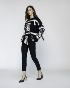 Vista completa de cuerpo entero del sweater negro con diseño gráfico blanco y detalles de flecos laterales, combinado con pantalón oscuro y stilettos